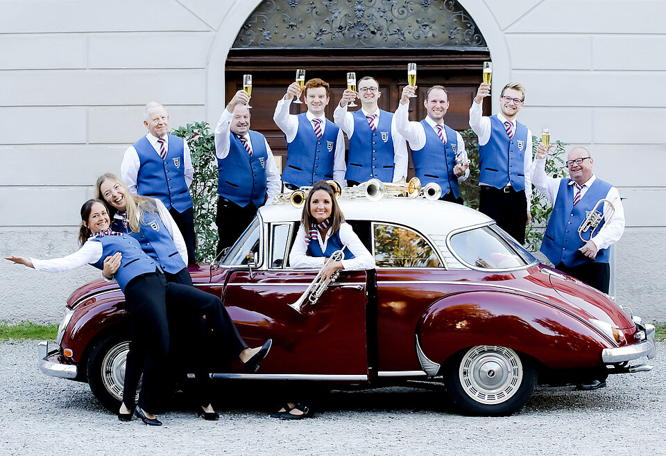 Musikverein Bad Waldsee Eine Gruppe von neun Menschen in blauen Westen und roten Krawatten posiert fröhlich um und auf einem roten Oldtimer-Auto, einige halten Musikinstrumente, andere heben Gläser zum Anstoßen.