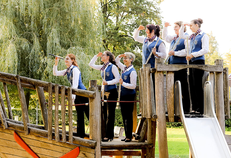 Holzblasregister Musikverein Bad Waldsee Sechs Frauen in einheitlicher Kleidung stehen auf einem hölzernen Spielplatz-Piratenschiff und schauen konzentriert in die Ferne, einige mit kleinen Fernrohren oder Händen über den Augen als Sonnenschutz.