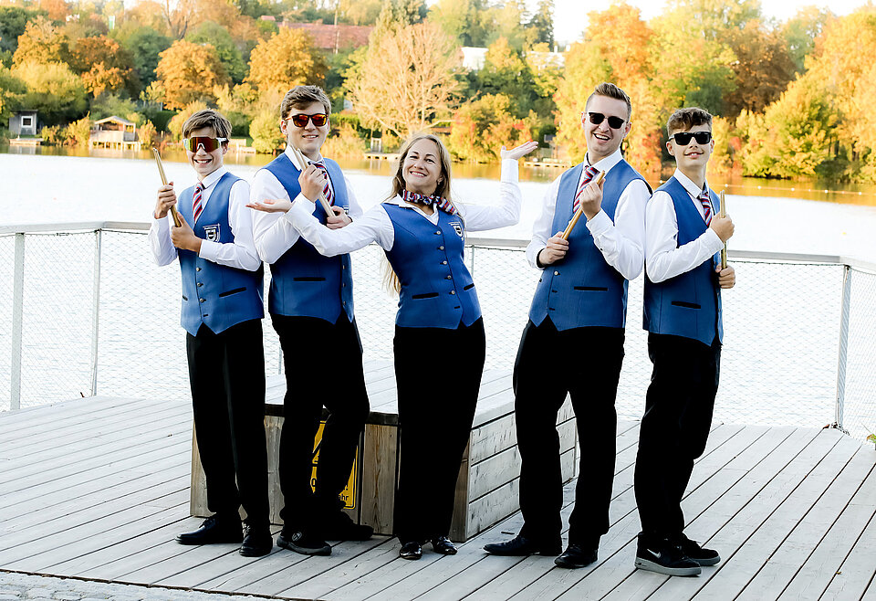 Musiker des Musikverein Bad Waldsee Fünf junge Musiker stehen auf einem Steg am Wasser, tragen blaue Westen und weiße Hemden, halten Trommelstöcke und lächeln in die Kamera.