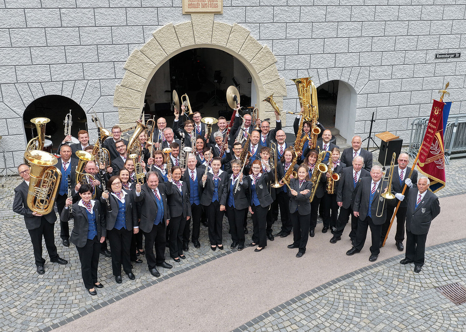 Eine große Blaskapelle mit über 40 Personen steht in einem Halbkreis vor einem Steinbogen. Die Musiker tragen schwarze Kleidung mit blauen Krawatten oder Schals und halten verschiedene Blasinstrumente wie Trompeten, Tuben und Posaunen.