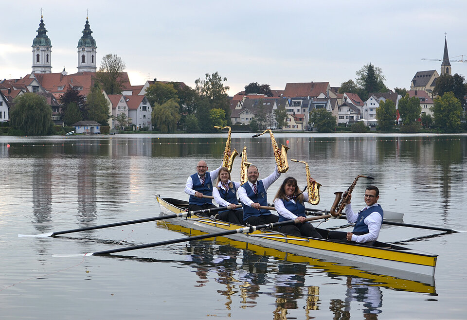 Musikverein Bad Waldsee Team Fünf Personen sitzen in einem Ruderboot auf einem See und halten Saxophone, im Hintergrund eine Stadt mit Kirchtürmen und Häusern.