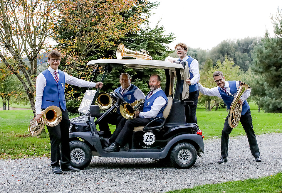 Tenorhörner Musikverein Bad Waldsee Fünf Musiker in blauen Westen mit Blasinstrumenten posieren fröhlich um und in einem kleinen Golfwagen auf einem Weg im Grünen.