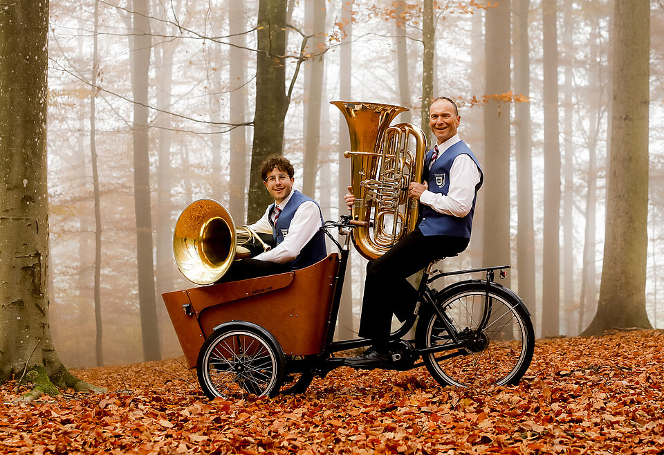 Tuba Musikverein Bad Waldsee Zwei Männer in Trachten sitzen in einem herbstlichen Wald auf einem Lastenfahrrad, einer spielt eine Tuba, der andere hält ein Sousafon im Fahrradkorb.