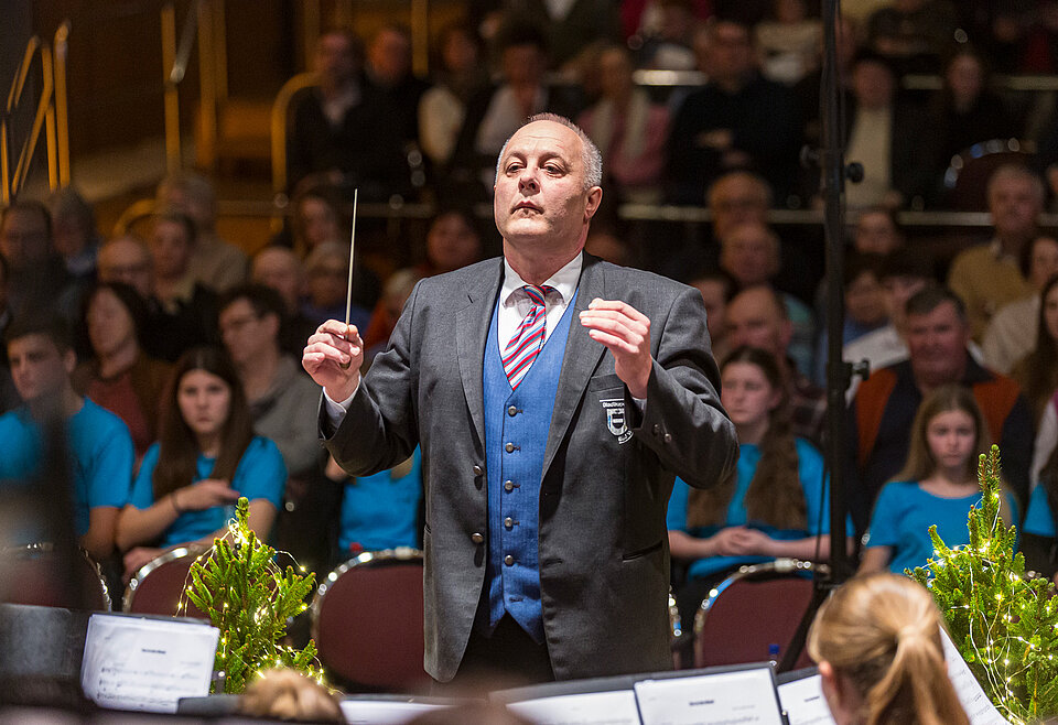 Dirigent Musikverein Bad Waldsee Ein Dirigent in formeller Kleidung leitet konzentriert ein Orchester vor einem Publikum in einem Konzertsaal.