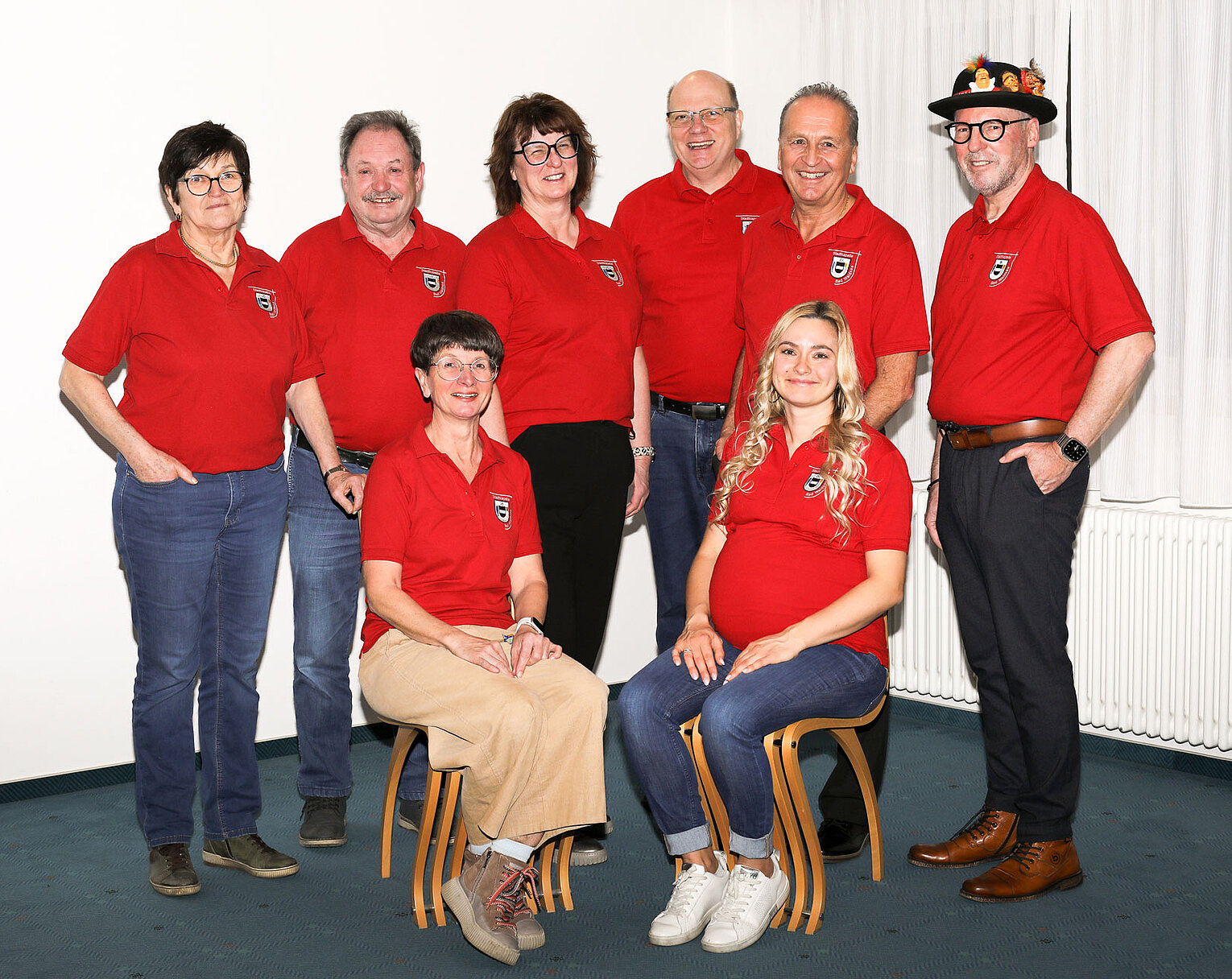 Acht Personen in roten Polo-Shirts posieren gemeinsam vor einem weißen Hintergrund, zwei Frauen sitzen vorn auf Stühlen, die übrigen fünf Personen stehen dahinter.