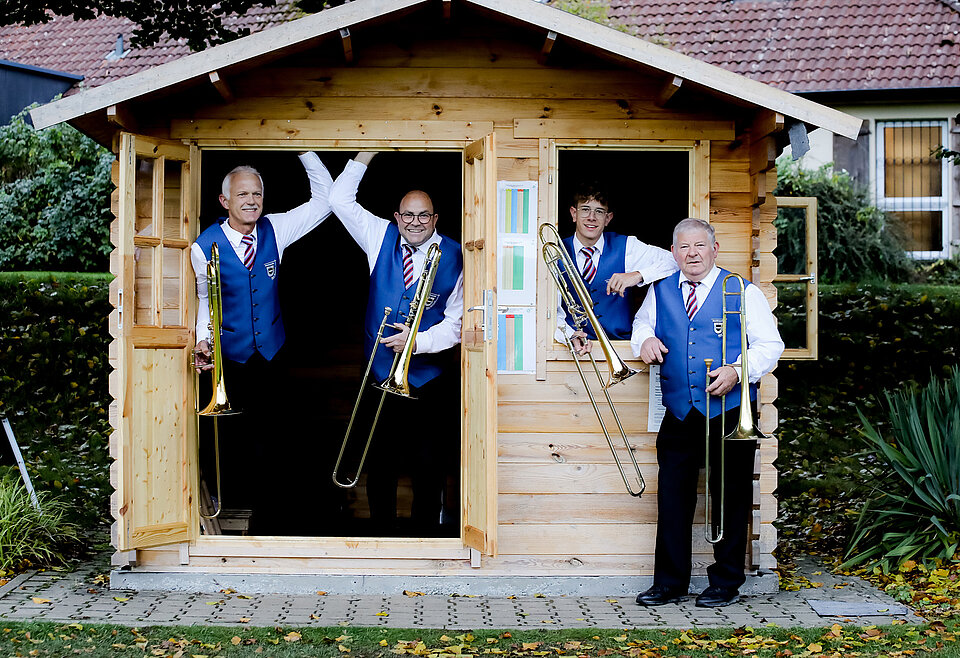 Posaunen Musikverein Bad Waldsee Vier ältere Männer in blauen Westen mit roten Krawatten stehen mit Posaunen vor und in einem kleinen Holzhaus und lächeln.